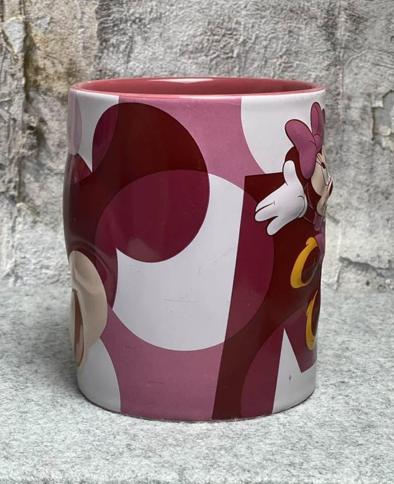 Op de afbeelding: Een cilindervormige keramische mok met een roze binnenkant en een buitenkant met een abstract cirkelpatroon in verschillende tinten roze en bordeauxrood. De mok heeft een reli&euml;f van een stripfiguur met een roze strik en witte handschoenen.