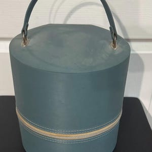 May include: A cylindrical, teal-colored handbag with a matching handle and a gold zipper. The bag has a rounded top and a base, with a zipper that runs around the middle. The handle is attached to the top with silver-colored metal rings.