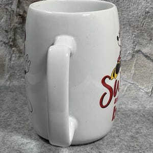 Pu&ograve; includere: Una tazza in ceramica bianca con la grafica di un personaggio dei cartoni animati con orecchie nere, un vestito rosso a pois bianchi e scarpe gialle. Il testo sulla tazza recita Sweet as can be in corsivo rosso. La tazza ha un manico curvo e finitura liscia.