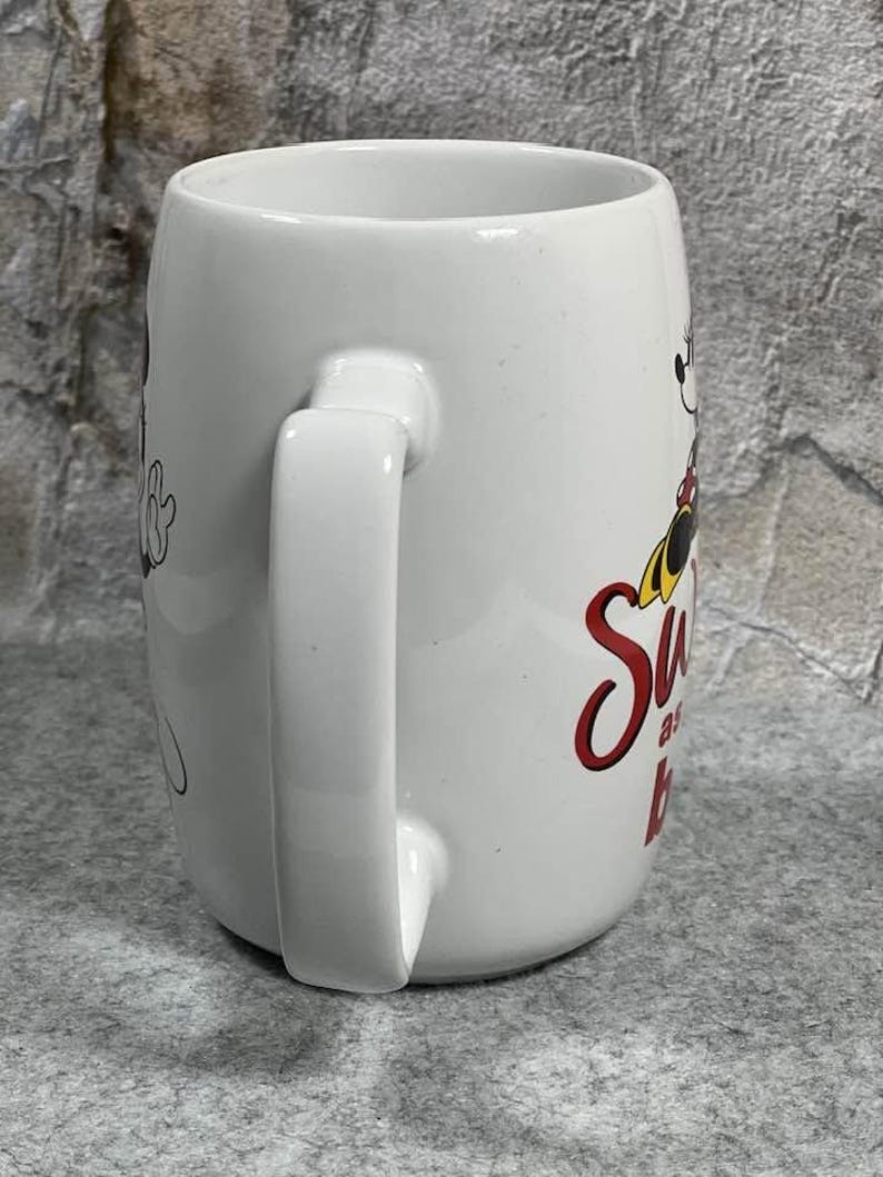 Pu&ograve; includere: Una tazza in ceramica bianca con la grafica di un personaggio dei cartoni animati con orecchie nere, un vestito rosso a pois bianchi e scarpe gialle. Il testo sulla tazza recita Sweet as can be in corsivo rosso. La tazza ha un manico curvo e finitura liscia.
