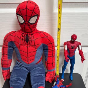 Puede incluir: Un mu&ntilde;eco grande de Spider-Man de peluche, de aproximadamente 40 cm de altura, con un disfraz rojo y azul. Tambi&eacute;n hay una figura de acci&oacute;n roja y azul y una figura de acci&oacute;n m&aacute;s peque&ntilde;a azul y roja. Una cinta m&eacute;trica amarilla est&aacute; al lado del mu&ntilde;eco.