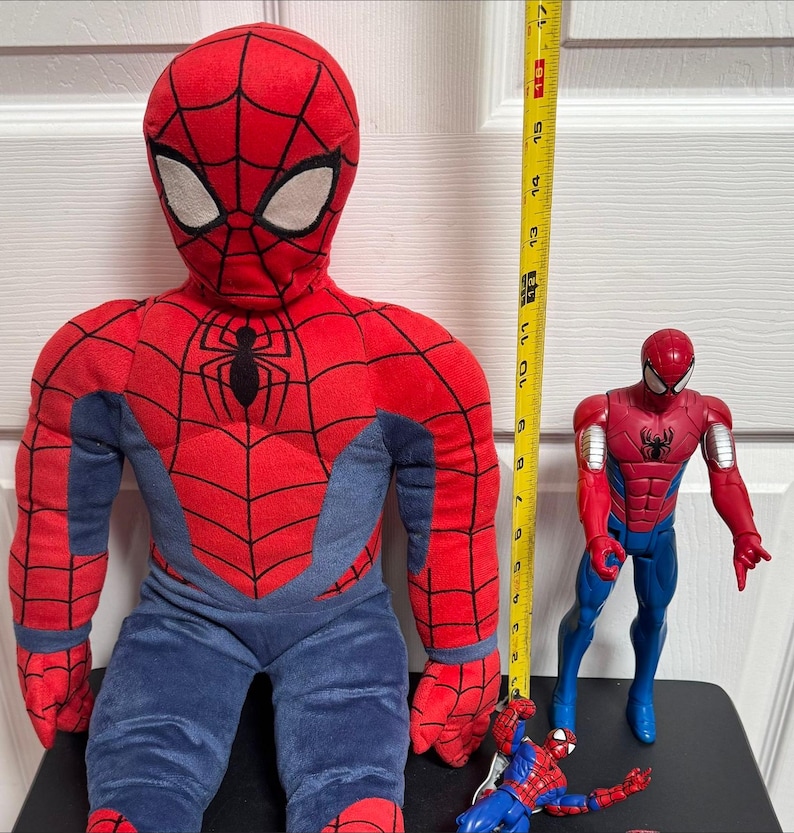 Puede incluir: Un mu&ntilde;eco grande de Spider-Man de peluche, de aproximadamente 40 cm de altura, con un disfraz rojo y azul. Tambi&eacute;n hay una figura de acci&oacute;n roja y azul y una figura de acci&oacute;n m&aacute;s peque&ntilde;a azul y roja. Una cinta m&eacute;trica amarilla est&aacute; al lado del mu&ntilde;eco.