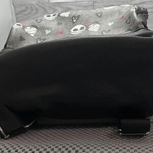 Puede incluir: Mochila negra con una parte superior estampada con caras de calaveras blancas y detalles en rosa. La parte inferior de la mochila es de color negro liso, con correas negras y hebillas plateadas. La mochila est&aacute; sobre una superficie gris.