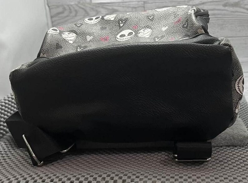 Puede incluir: Mochila negra con una parte superior estampada con caras de calaveras blancas y detalles en rosa. La parte inferior de la mochila es de color negro liso, con correas negras y hebillas plateadas. La mochila est&aacute; sobre una superficie gris.