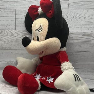 Puede incluir: Una mu&ntilde;eca de peluche de Minnie Mouse con un atuendo festivo. La mu&ntilde;eca tiene cabeza y orejas negras, cara color crema y un lazo rojo. Lleva un vestido rojo con detalles de copos de nieve blancos y guantes blancos. Las piernas y los pies de la mu&ntilde;eca son rojos.