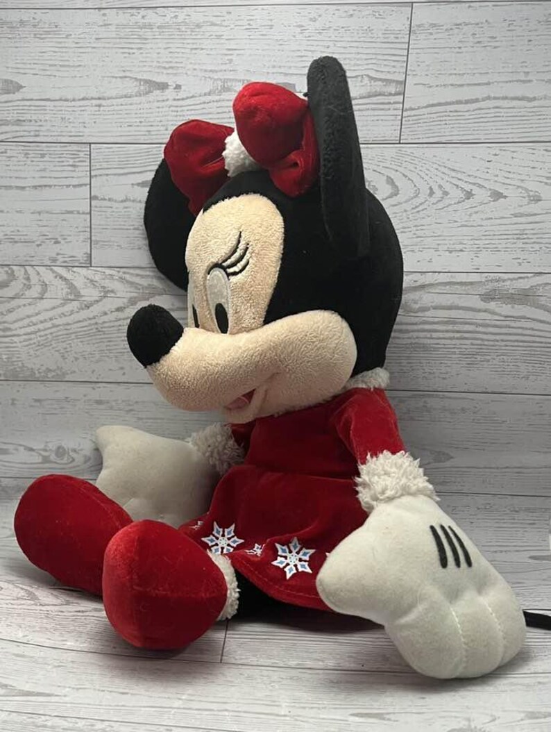 Puede incluir: Una mu&ntilde;eca de peluche de Minnie Mouse con un atuendo festivo. La mu&ntilde;eca tiene cabeza y orejas negras, cara color crema y un lazo rojo. Lleva un vestido rojo con detalles de copos de nieve blancos y guantes blancos. Las piernas y los pies de la mu&ntilde;eca son rojos.