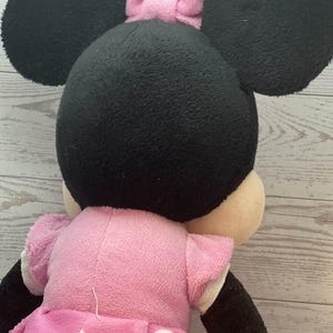 Pode incluir: Uma boneca de pel&uacute;cia da Minnie Mouse, vista por tr&aacute;s, com grandes orelhas pretas e um la&ccedil;o rosa. A boneca usa um vestido rosa com bolinhas brancas e mangas curtas. Os bra&ccedil;os e as pernas s&atilde;o pretos e brancos.