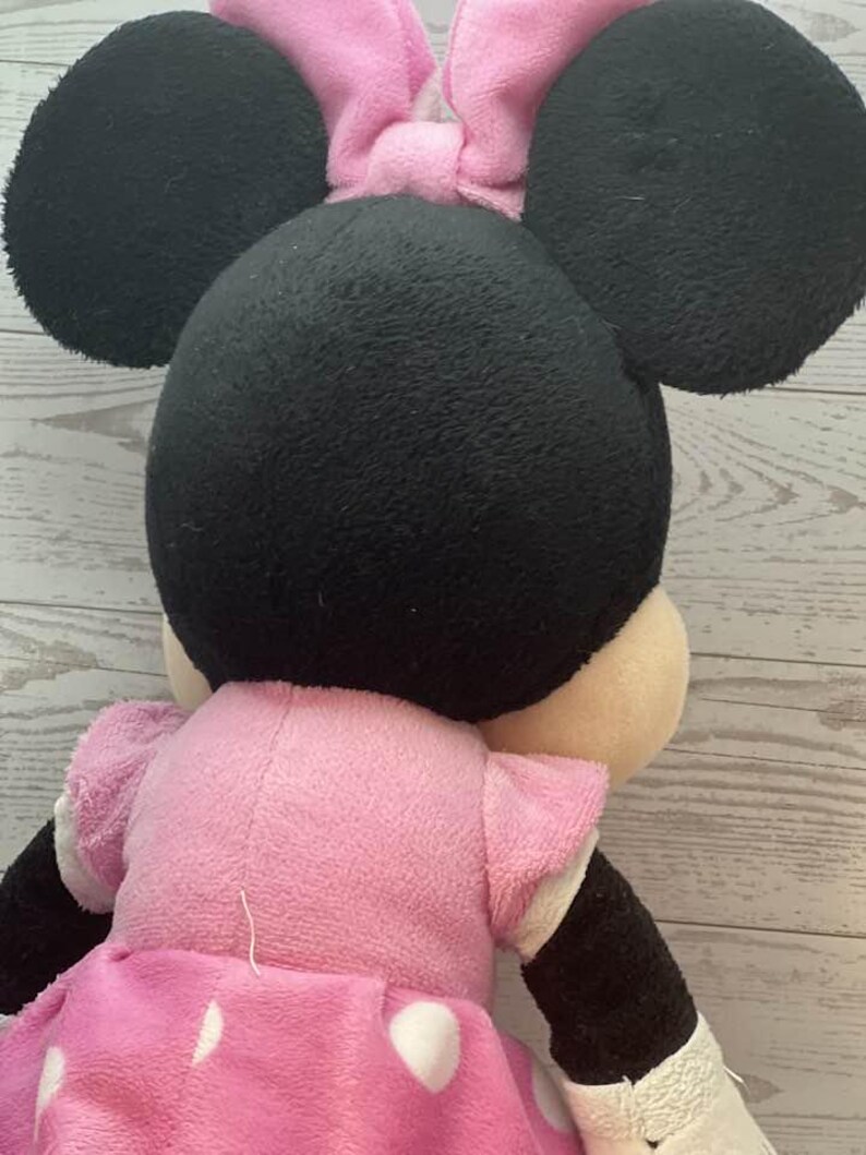 Pode incluir: Uma boneca de pel&uacute;cia da Minnie Mouse, vista por tr&aacute;s, com grandes orelhas pretas e um la&ccedil;o rosa. A boneca usa um vestido rosa com bolinhas brancas e mangas curtas. Os bra&ccedil;os e as pernas s&atilde;o pretos e brancos.