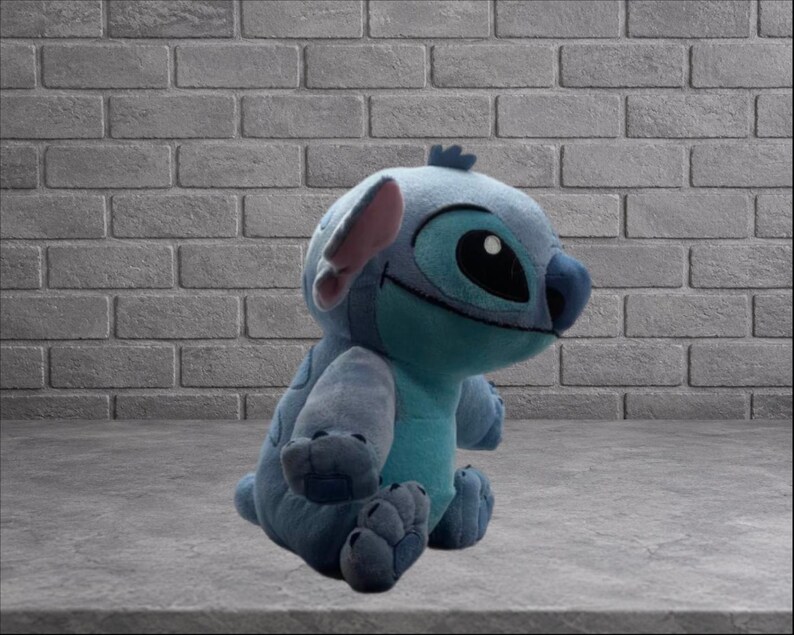 Puede incluir: Un peluche del personaje Stitch de la pel&iacute;cula de Disney Lilo & Stitch. El juguete es principalmente azul con una panza turquesa, ojos grandes negros y orejas internas rosas. Est&aacute; sentado sobre una superficie gris frente a una pared de ladrillos.
