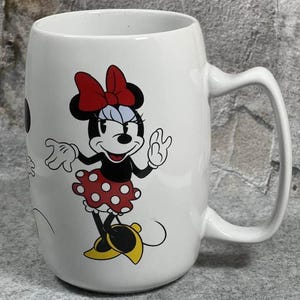 Pu&ograve; includere: Una tazza in ceramica bianca con un'illustrazione grafica di Minnie Mouse. Il personaggio indossa un fiocco rosso, un vestito rosso a pois bianchi e scarpe gialle. La tazza ha un corpo cilindrico liscio e un manico curvo sul lato destro.