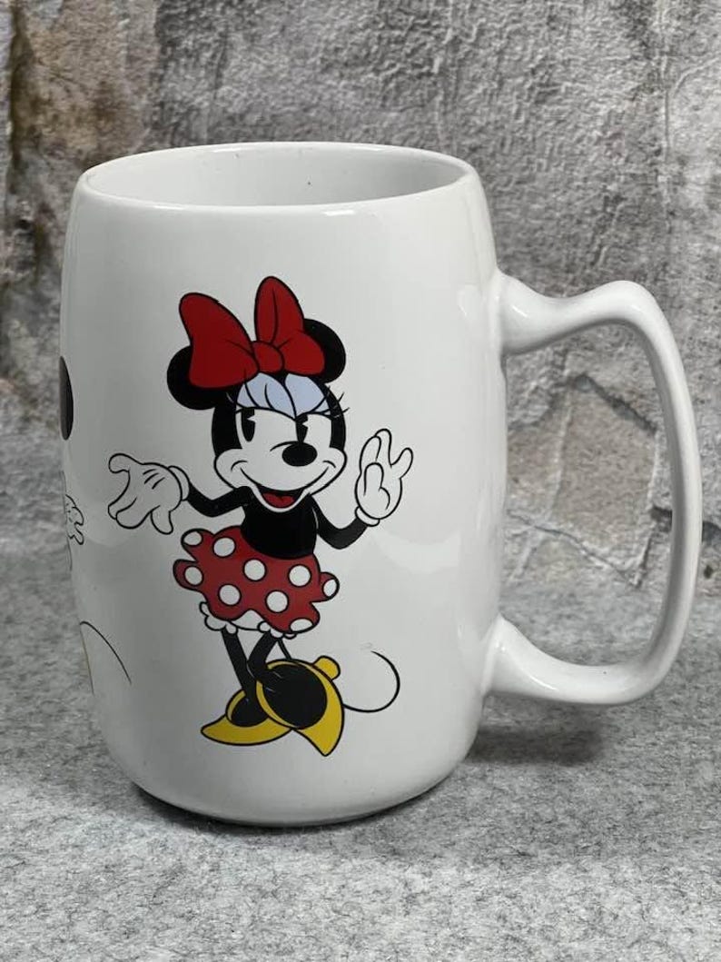 Pu&ograve; includere: Una tazza in ceramica bianca con un'illustrazione grafica di Minnie Mouse. Il personaggio indossa un fiocco rosso, un vestito rosso a pois bianchi e scarpe gialle. La tazza ha un corpo cilindrico liscio e un manico curvo sul lato destro.