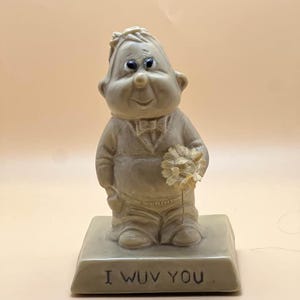 May include: A beige figurine of a smiling, cartoonish man holding a bouquet of flowers. The figurine is standing on a rectangular base with the words "I WUV YOU" in black lettering.