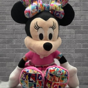 May include: A plush Minnie Mouse doll with a pink dress, black ears, and a colorful bow. The doll's feet feature a patchwork design with cartoon characters. The doll is sitting in front of a gray brick wall.