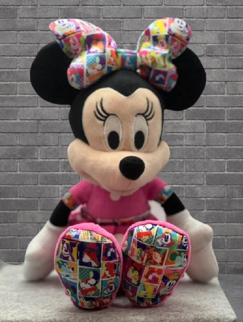May include: A plush Minnie Mouse doll with a pink dress, black ears, and a colorful bow. The doll's feet feature a patchwork design with cartoon characters. The doll is sitting in front of a gray brick wall.