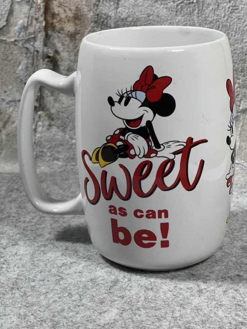Pu&ograve; includere: Una tazza in ceramica bianca con un'illustrazione di Minnie Mouse che indossa un fiocco rosso e un vestito a pois. Il testo "Sweet as can be!" &egrave; stampato in rosso, in corsivo e in stampatello, sotto il personaggio. La tazza ha un manico curvo.