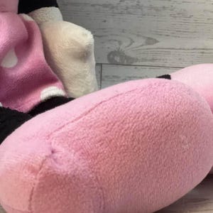 Pode incluir: Um brinquedo de pel&uacute;cia com um p&eacute; e uma perna rosa. O p&eacute; &eacute; rosa texturizado, com costuras vis&iacute;veis. O fundo inclui uma superf&iacute;cie de madeira branca e cinza. O brinquedo faz parte de um personagem maior.
