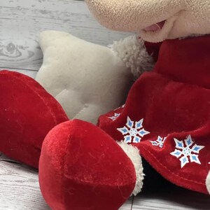 Puede incluir: Un mu&ntilde;eco de peluche de Mickey Mouse con un atuendo de terciopelo rojo con detalles y adornos de copos de nieve blancos. El mu&ntilde;eco tiene cabeza negra, guantes blancos y pies de terciopelo rojo. Fondo de superficie de madera clara.