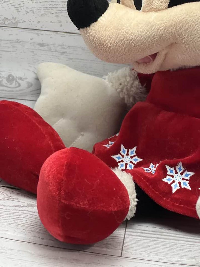 Puede incluir: Un mu&ntilde;eco de peluche de Mickey Mouse con un atuendo de terciopelo rojo con detalles y adornos de copos de nieve blancos. El mu&ntilde;eco tiene cabeza negra, guantes blancos y pies de terciopelo rojo. Fondo de superficie de madera clara.