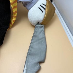 May include: A plush toy hand with a white glove, gold cuff, and three black lines. The hand is attached to a gray, metallic-looking blade. The sleeve is gray and the shirt is yellow and orange striped.
