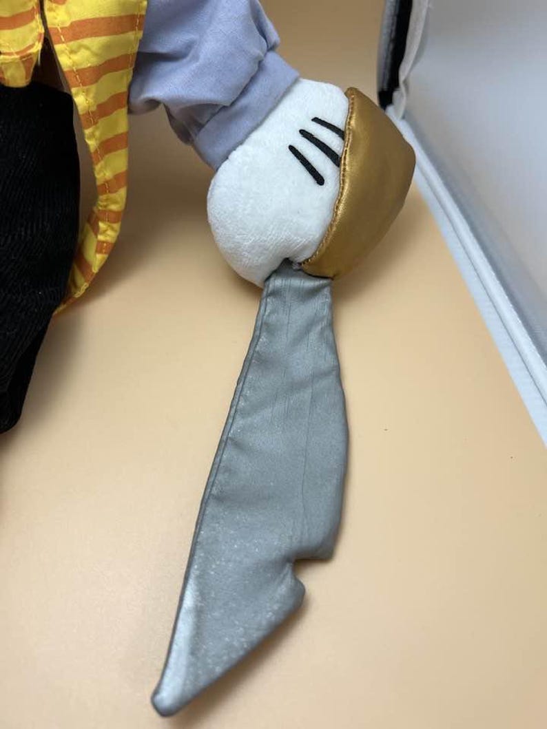 May include: A plush toy hand with a white glove, gold cuff, and three black lines. The hand is attached to a gray, metallic-looking blade. The sleeve is gray and the shirt is yellow and orange striped.