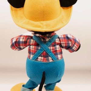 May include: A plush toy of a character wearing a yellow hat with black ears, a red, white, and blue plaid long-sleeve shirt, and blue overalls with crisscross straps. The character has a black tail and large yellow shoes with light blue cuffs.
