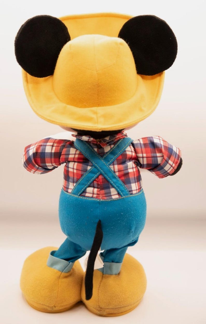 May include: A plush toy of a character wearing a yellow hat with black ears, a red, white, and blue plaid long-sleeve shirt, and blue overalls with crisscross straps. The character has a black tail and large yellow shoes with light blue cuffs.