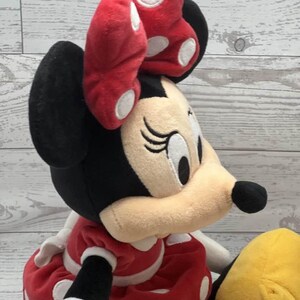 May include: A Minnie Mouse soft toy featuring a red bow with white spots. The doll has black ears and head, a beige face, and a red dress with white spots. A yellow plush is also visible.