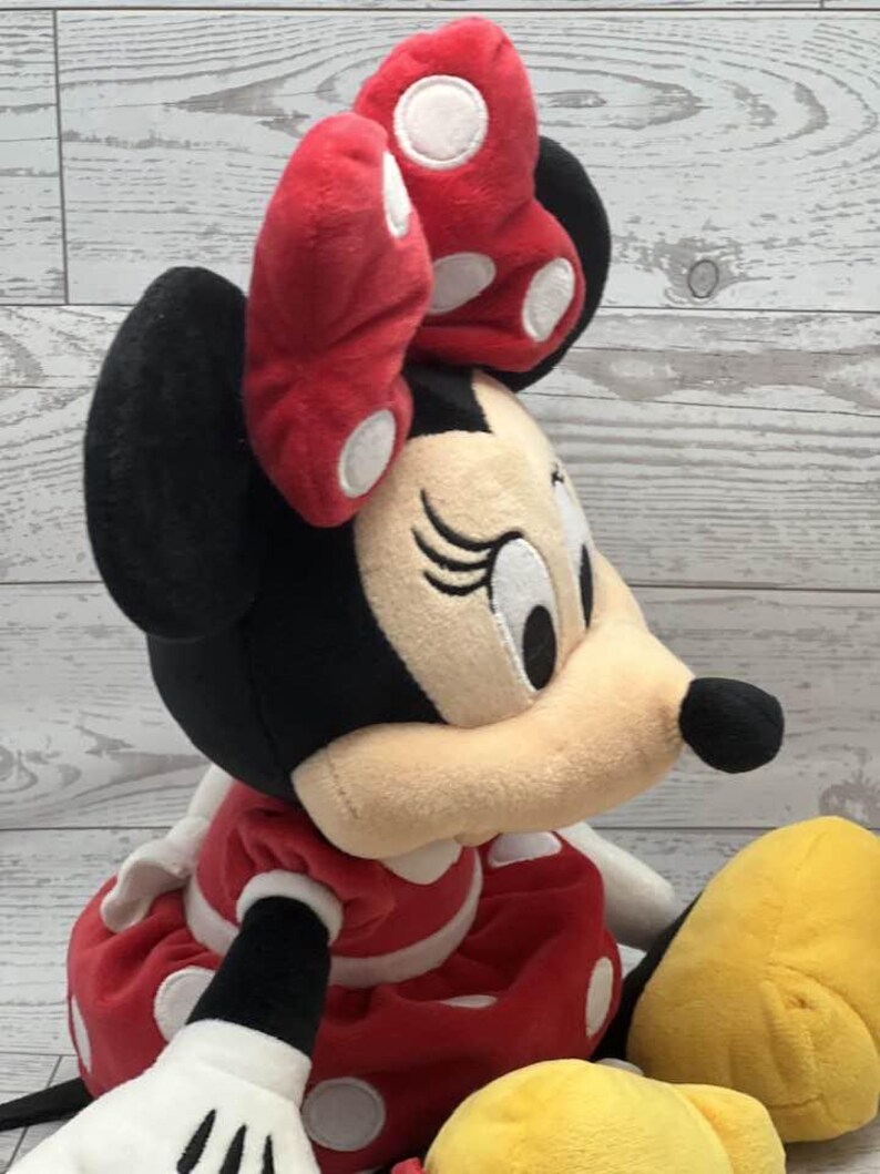 May include: A Minnie Mouse soft toy featuring a red bow with white spots. The doll has black ears and head, a beige face, and a red dress with white spots. A yellow plush is also visible.