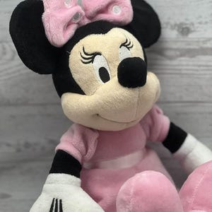 May include: A plush Minnie Mouse doll, featuring a pink dress, bow, and signature black ears. The doll has white gloves with black stripes and closed eyes, creating a classic and recognizable character.
