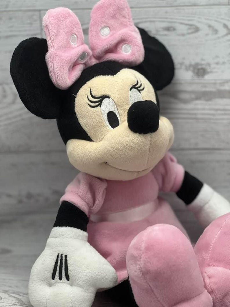May include: A plush Minnie Mouse doll, featuring a pink dress, bow, and signature black ears. The doll has white gloves with black stripes and closed eyes, creating a classic and recognizable character.
