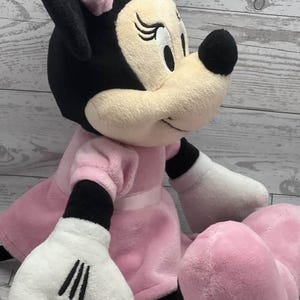 May include: A plush Minnie Mouse doll, featuring a black head and ears, a pink bow, and a pink dress. The doll has white gloves and pink feet. The doll is sitting down and has a friendly expression.