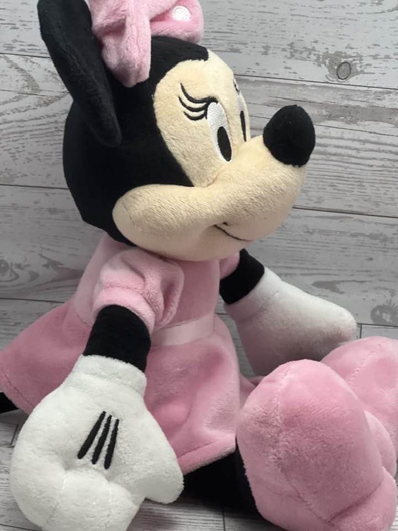 May include: A plush Minnie Mouse doll, featuring a black head and ears, a pink bow, and a pink dress. The doll has white gloves and pink feet. The doll is sitting down and has a friendly expression.