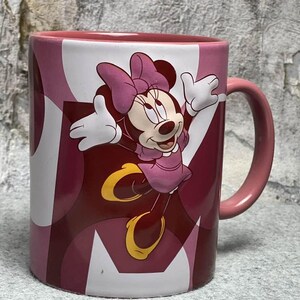 Op de afbeelding: Een keramische mok met een afbeelding van Minnie Mouse met haar armen omhoog, gekleed in een roze jurk en strik. De mok heeft een roze handvat en een achtergrondpatroon van overlappende cirkels in tinten roze, donkerrood en wit.