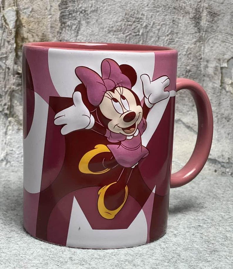 Op de afbeelding: Een keramische mok met een afbeelding van Minnie Mouse met haar armen omhoog, gekleed in een roze jurk en strik. De mok heeft een roze handvat en een achtergrondpatroon van overlappende cirkels in tinten roze, donkerrood en wit.