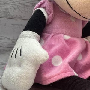 Pode incluir: Uma boneca de pel&uacute;cia da Minnie Mouse, com um vestido rosa com bolinhas brancas, uma luva branca com linhas pretas e bra&ccedil;os e pernas pretas. A boneca tem um rosto sorridente e um la&ccedil;o preto.