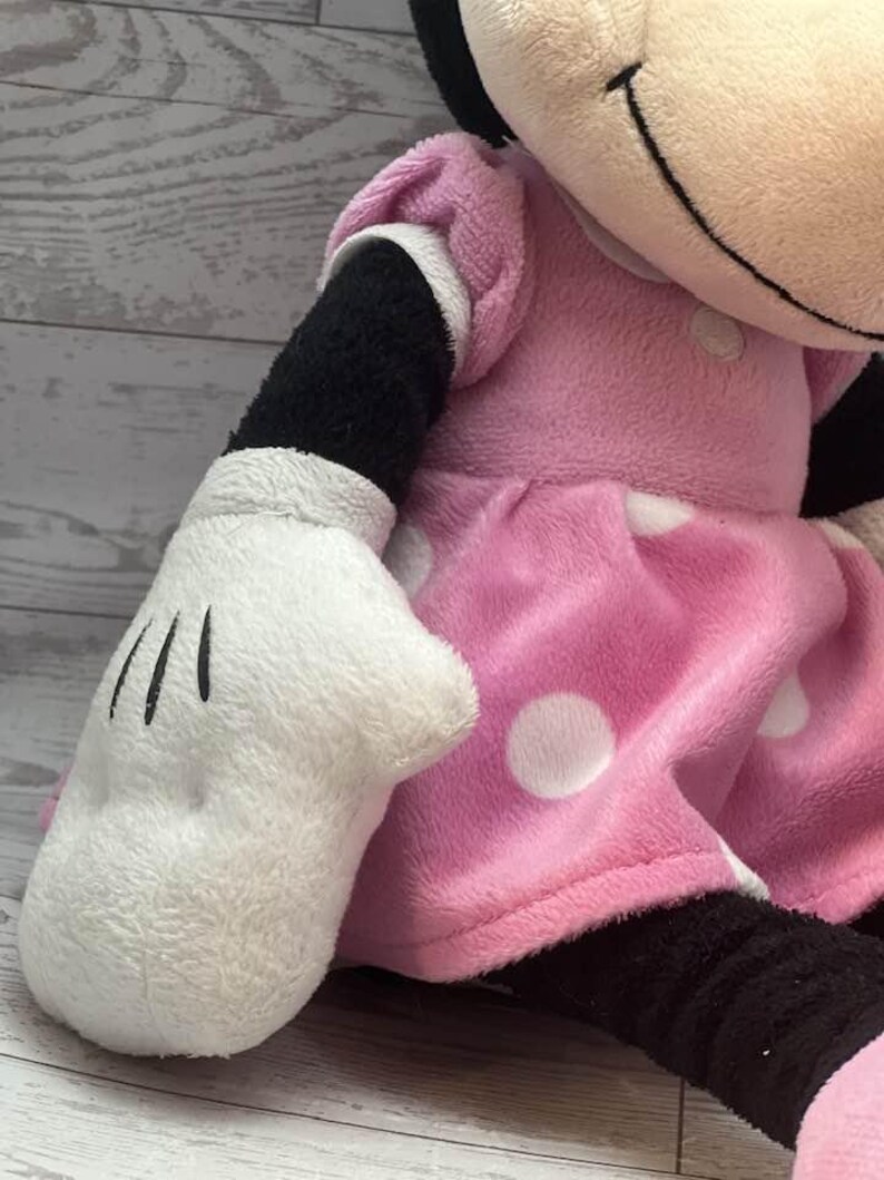 Pode incluir: Uma boneca de pel&uacute;cia da Minnie Mouse, com um vestido rosa com bolinhas brancas, uma luva branca com linhas pretas e bra&ccedil;os e pernas pretas. A boneca tem um rosto sorridente e um la&ccedil;o preto.