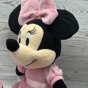 May include: A plush Minnie Mouse doll, featuring a pink bow with white polka dots, black ears, and a pink dress. The doll has a cream-colored face, black nose, and closed eyes. The doll's arms and legs are black and white.