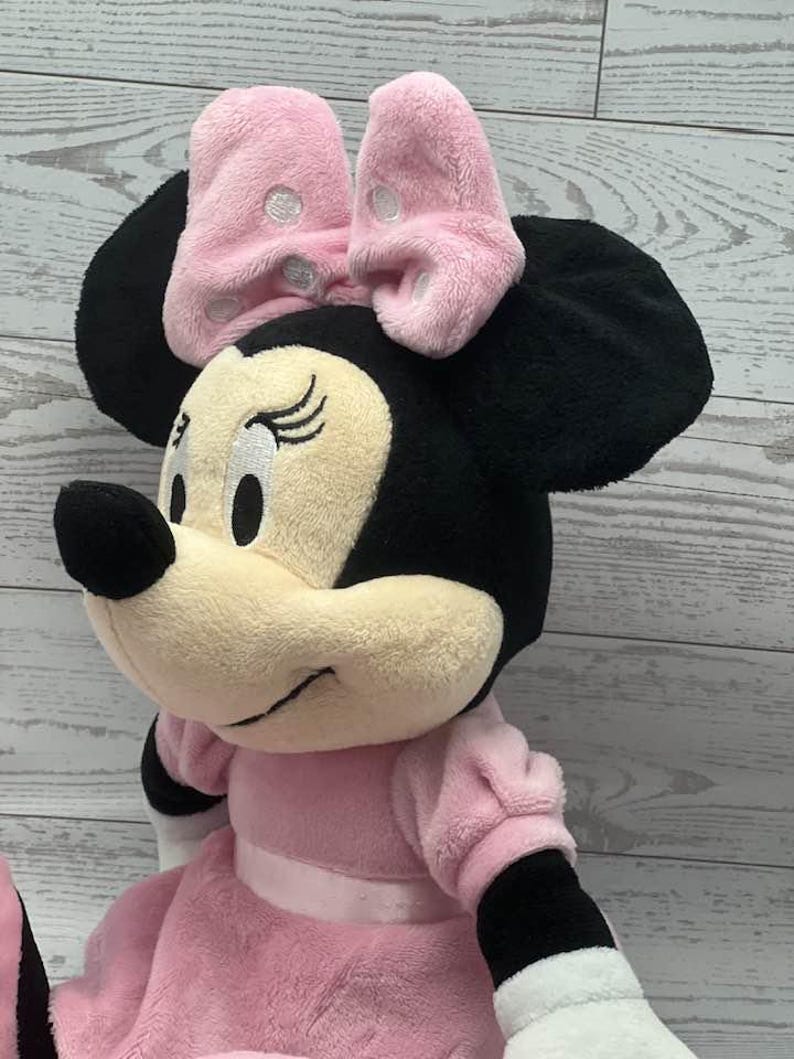 May include: A plush Minnie Mouse doll, featuring a pink bow with white polka dots, black ears, and a pink dress. The doll has a cream-colored face, black nose, and closed eyes. The doll's arms and legs are black and white.