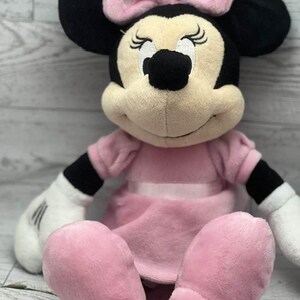 May include: A plush Minnie Mouse doll, featuring a pink dress and bow with white polka dots. The doll has black ears, a tan face, and white gloves. The doll is sitting with its legs extended.