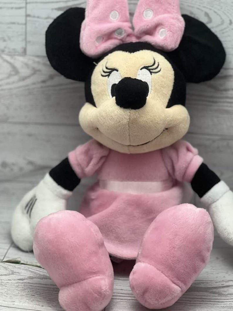 May include: A plush Minnie Mouse doll, featuring a pink dress and bow with white polka dots. The doll has black ears, a tan face, and white gloves. The doll is sitting with its legs extended.