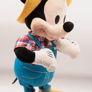 May include: A plush Mickey Mouse figure wearing a yellow hat, a red and blue plaid shirt, and blue overalls with yellow stitching. The character has white gloves, large yellow shoes, and a black head with round ears, standing in a side profile view.