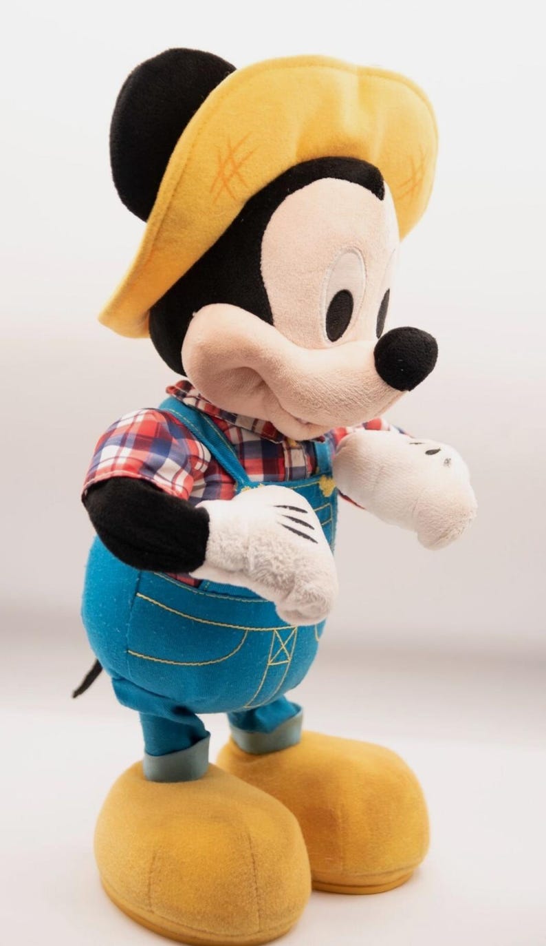 May include: A plush Mickey Mouse figure wearing a yellow hat, a red and blue plaid shirt, and blue overalls with yellow stitching. The character has white gloves, large yellow shoes, and a black head with round ears, standing in a side profile view.