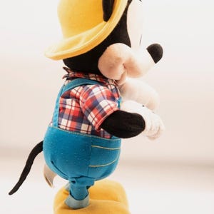 May include: A plush Mickey Mouse toy wearing a yellow hat, a red and white plaid shirt, and blue denim-style overalls with yellow stitching. The character has black ears, a white face, white gloves, and large yellow shoes, standing in profile.