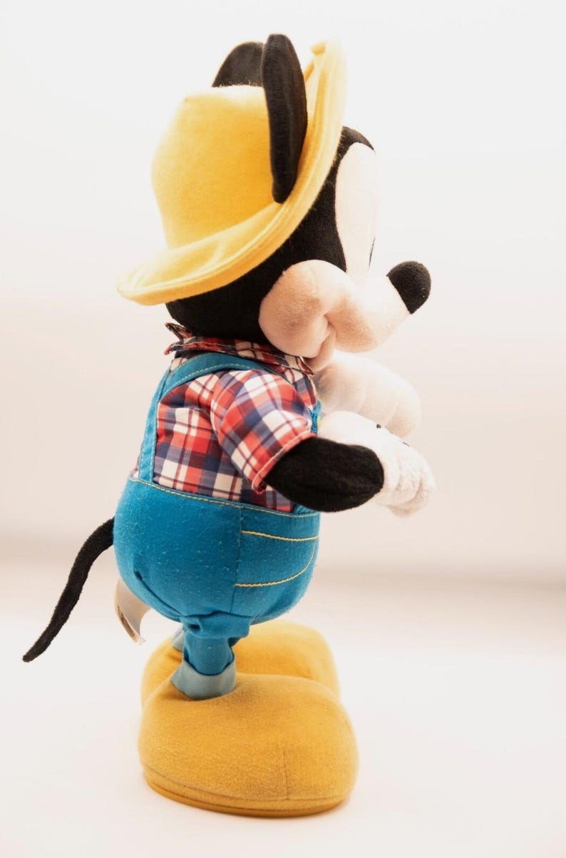 May include: A plush Mickey Mouse toy wearing a yellow hat, a red and white plaid shirt, and blue denim-style overalls with yellow stitching. The character has black ears, a white face, white gloves, and large yellow shoes, standing in profile.