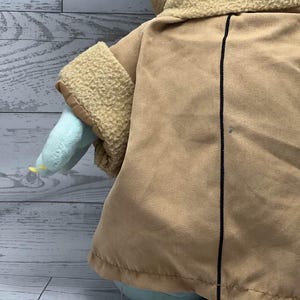 May include: A plush toy of a character with large ears, light green skin, and a tan coat. The coat has a black vertical line down the centre. The toy's sleeves are tan with a textured trim. The toy is on a light grey wooden surface.