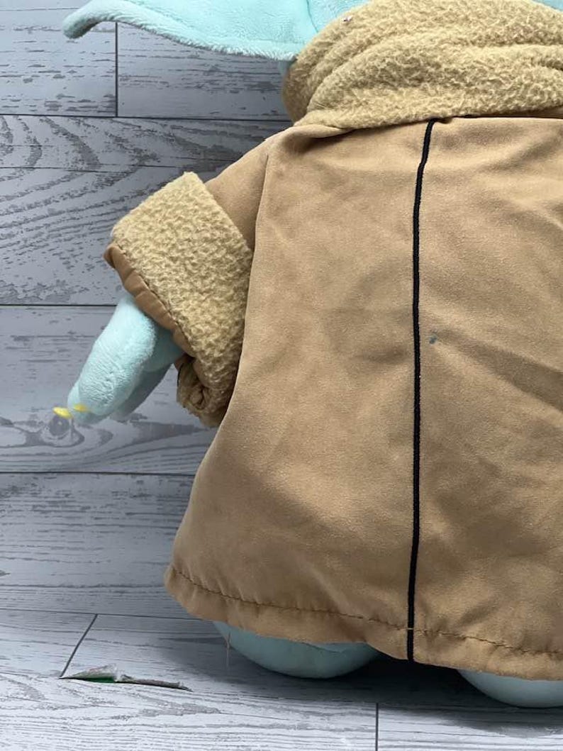 May include: A plush toy of a character with large ears, light green skin, and a tan coat. The coat has a black vertical line down the centre. The toy's sleeves are tan with a textured trim. The toy is on a light grey wooden surface.