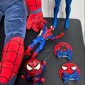 Puede incluir: Una colecci&oacute;n de art&iacute;culos con tem&aacute;tica de Spider-Man. Incluye una pierna y un brazo de peluche, dos figuras de acci&oacute;n y dos adornos de pajarita de lentejuelas rojas y azules. Las figuras y los adornos est&aacute;n sobre una superficie negra.
