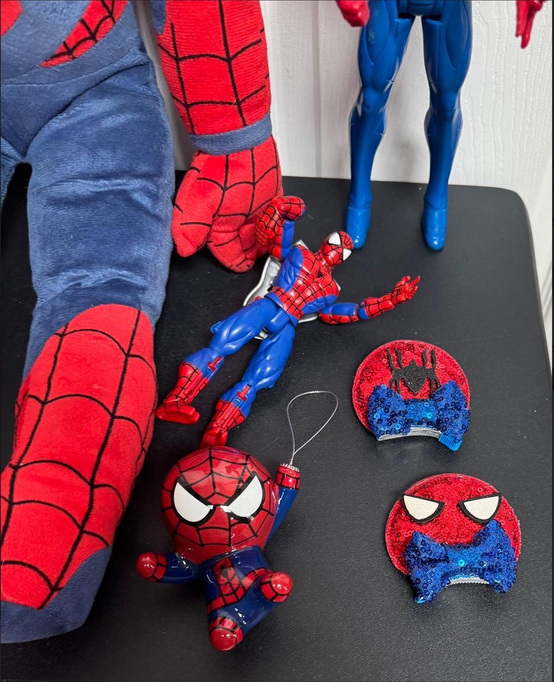Puede incluir: Una colecci&oacute;n de art&iacute;culos con tem&aacute;tica de Spider-Man. Incluye una pierna y un brazo de peluche, dos figuras de acci&oacute;n y dos adornos de pajarita de lentejuelas rojas y azules. Las figuras y los adornos est&aacute;n sobre una superficie negra.