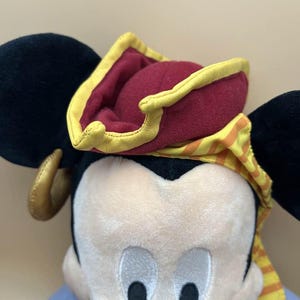 May include: A plush toy of a cartoon mouse wearing a pirate hat. The hat is burgundy with a yellow trim and a yellow and orange striped scarf. The mouse has black ears and a light-colored face with black eyes.