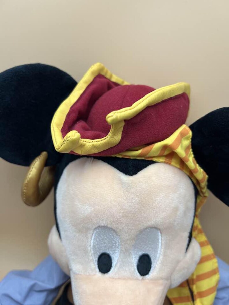 May include: A plush toy of a cartoon mouse wearing a pirate hat. The hat is burgundy with a yellow trim and a yellow and orange striped scarf. The mouse has black ears and a light-colored face with black eyes.